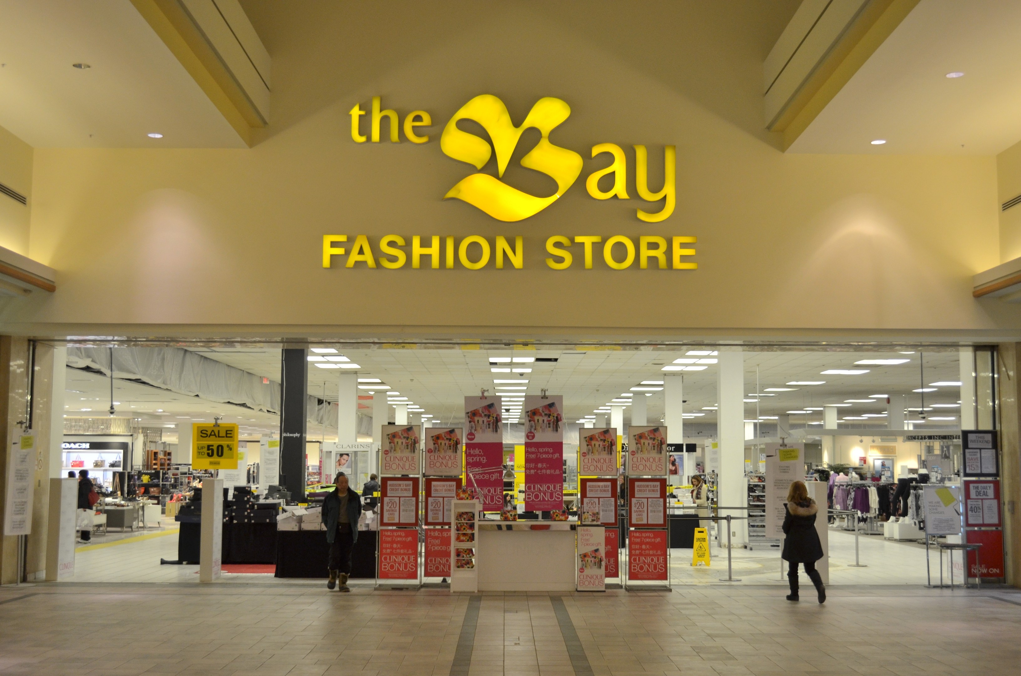 Außenansicht eines Bay Fashion Stores in einem Einkaufszentrum mit Menschen, die gehen, Texttafeln an den Wänden, Säulen, Deckenleuchten und Hintergrundobjekte.