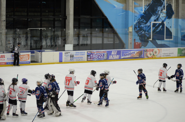 Gruppe junger Menschen, die Eisockey auf einer Indoor-Eisfläche spielen, mit Helmen und Stöcken, sowie einer Person links daneben und Bannern, Säulen und Glaswänden im Hintergrund.