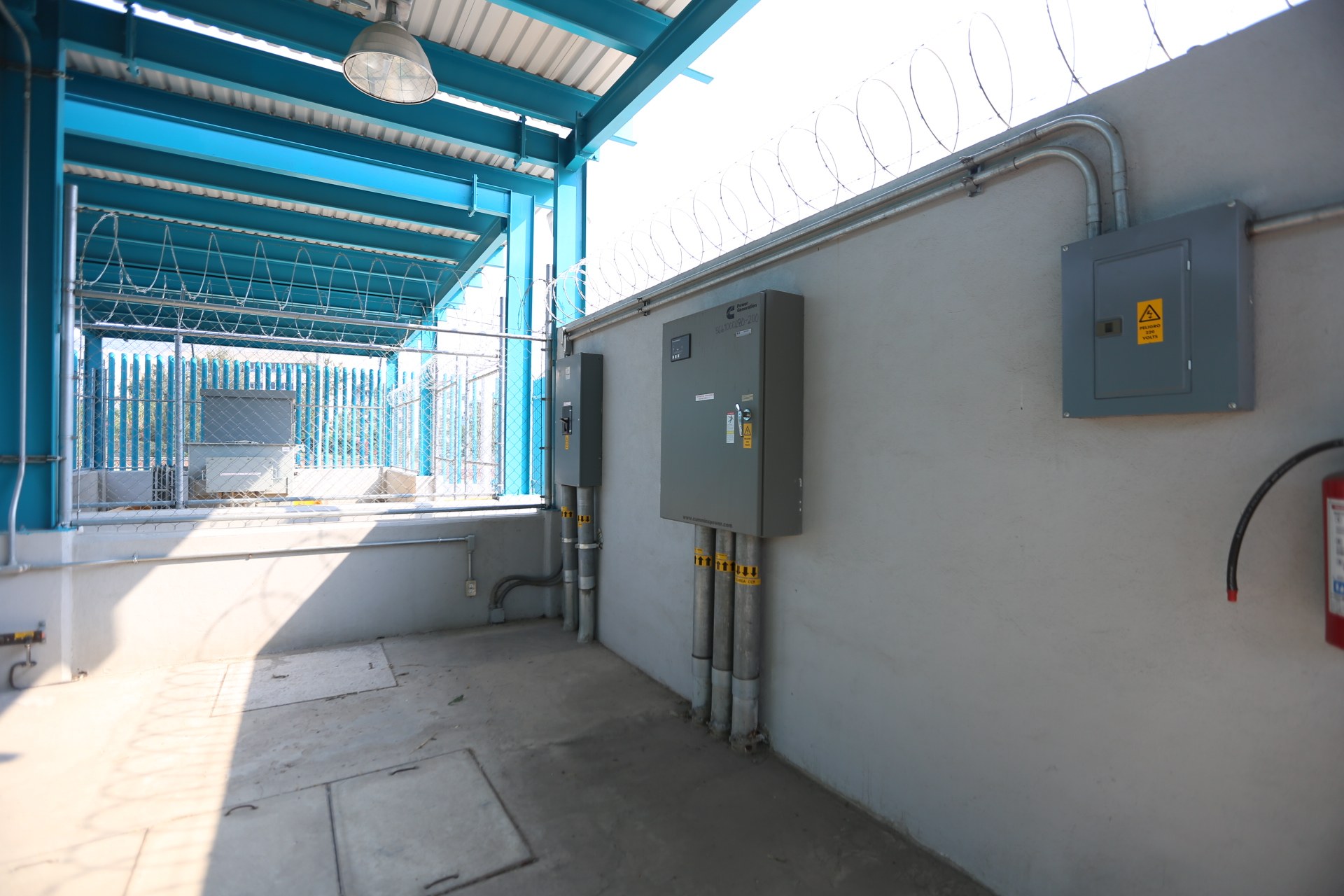 Raum mit einem roten Feuerlöscher auf der rechten Seite, elektrischen Schaltkästen und Rohren an der Wand, einer Deckenleuchte und einem Zaun im Hintergrund, was auf eine elektrische Umspannstation hindeutet.