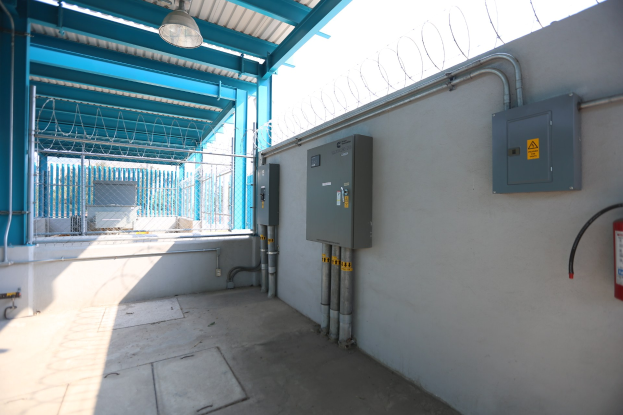 Raum mit einem roten Feuerlöscher auf der rechten Seite, elektrischen Schaltkästen und Rohren an der Wand, einer Deckenleuchte und einem Zaun im Hintergrund, was auf eine elektrische Umspannstation hindeutet.