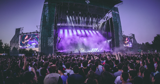 Eine große Menschenmenge bei einem Musikfestival schaut auf einer beleuchteten Bühne mit Instrumenten und Bildschirmen, die vor einem Gebäude, Bäumen und einem klaren blauen Himmel steht.