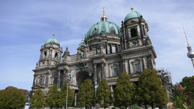 Der Berliner Dom, eine große Kathedrale mit Fenstern, Säulen, Bögen, Statuen und einem Turm, steht prominent in Berlin, Deutschland, mit Menschen in der Nähe, Bäumen, Blumenpflanzen und einem bewölkten Himmel im Hintergrund.
