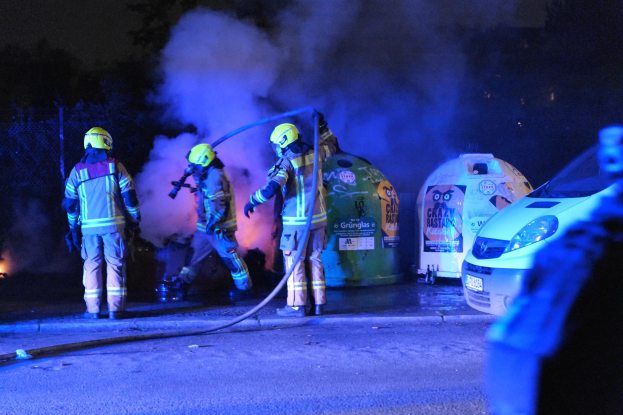 Feuerwehrleute in Helmen auf einer Straße stehend, Höschenschläuche haltend, vor einem Auto, das in Flammen steht, mit Rauchwolken, Bäumen und einem Zaun im Hintergrund.