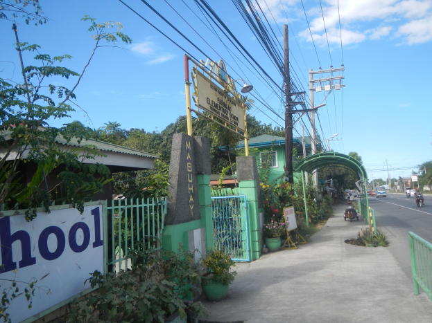 Schuleingangsschild am Straßenrand mit einem Bogen, Tor und Namenskennzeichen, umgeben von Fahrzeugen, Pflanzen, Bäumen, Strommästen und einem bewölkten Himmel.