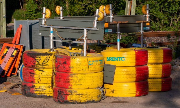 Ein Haufen gelber und roter Reifen auf einem Schmutzfeld umgeben von verstreuten Metallobjekten, Brettern und anderem Schutt, mit Bäumen, Pflanzen und einem Schuppen im Hintergrund.