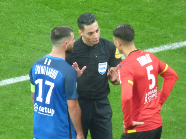 Schiedsrichter in schwarzem Hemd und Hose, der mit zwei Männern in Brille auf einem Fußballfeld spricht, einer trägt ein blau-rotes Hemd mit Text.