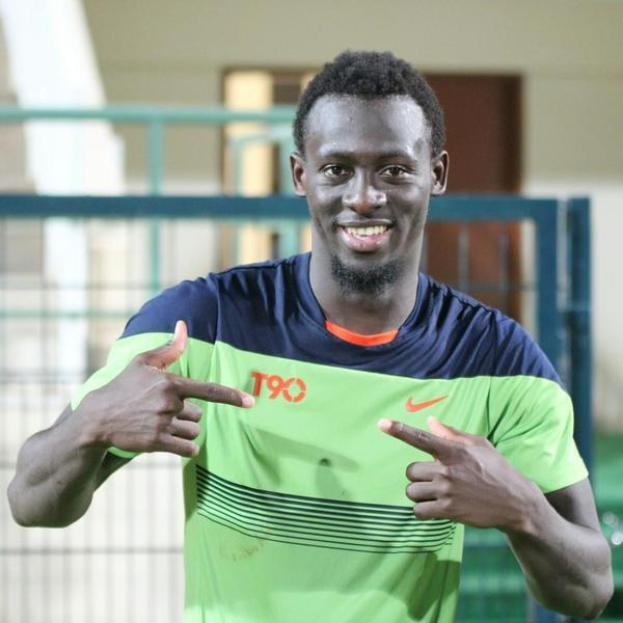 Ein Mann in einem grünen und blauen Hemd, der in die Kamera lächelt und zeigt, mit einem unscharfen Hintergrund, der eine Brüstung, eine Wand und eine Tür zeigt.