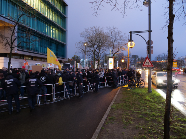 Große Gruppe von Menschen protestiert vor einem Gebäude in Berlin, hält Schilder, mit Barrikaden, Fahrrädern, Laternenpfählen, Schildern, Bäumen, Gras und Himmel im Hintergrund.