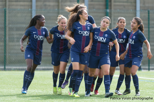 Gruppe junger Frauen beim Fußballspielen auf einem Rasenfeld mit Maschendrahtzaun und einer Wand im Hintergrund, Text in der rechten unteren Ecke lautet "Paris Saint-Germain Women's Soccer".