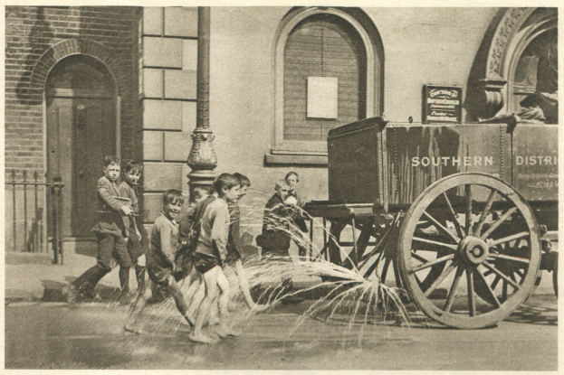 Schwarz-weißes Bild von Kindern, die mit einem Hydranten vor einem Gebäude mit Fenstern, einer Tür, einem Mast und einem Zaun spielen, mit einem Wagen im Hintergrund.