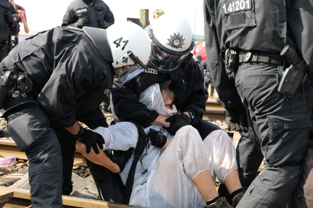 Eine Gruppe von Polizisten in Einsatzausrüstung verhaftet einen Mann in einem weißen Kleid auf einem Bahngleis, während andere im Hintergrund stehen.