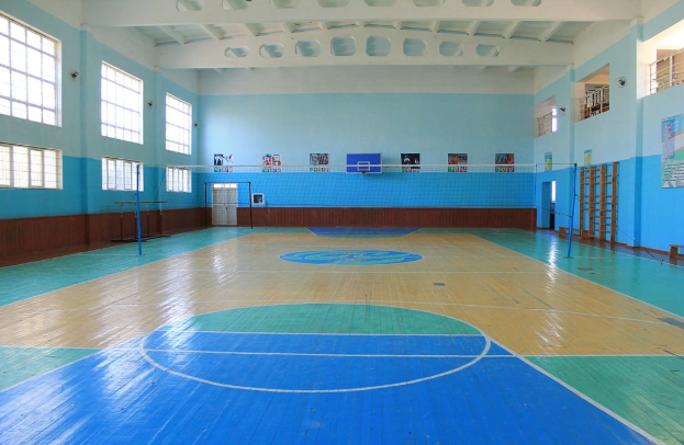 Große Turnhalle mit einem blauen und weißen Basketballfeld, einem Netz an Pfosten, einer Tür, Fenstern, Plakaten an den Wänden, einem Geländer und einem Dach.