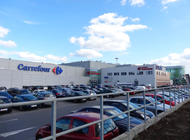 Großes Autohaus mit zahlreichen parkenden Fahrzeugen vorne, umgeben von städtischen Gebäuden, Straßeninfrastruktur und einem bewölkten Himmel.