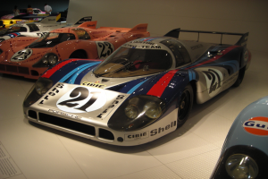 Ein Porsche 917K Le Mans Rennwagen wird in einem Museum ausgestellt, umgeben von anderen Sportwagen und Menschen, mit einer Wand im Hintergrund und einem Blatt Papier mit Text links.