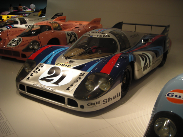 Ein Porsche 917K Le Mans Rennwagen wird in einem Museum ausgestellt, umgeben von anderen Sportwagen und Menschen, mit einer Wand im Hintergrund und einem Blatt Papier mit Text links.