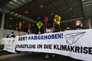 Eine Gruppe maskierter Personen mit einem Banner mit der Aufschrift "Sturzflug in die Klimakrise" steht vor einem Glasgebäude mit Säulen, beleuchtet von Deckenlampen.