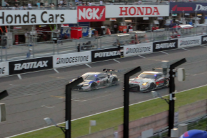 Eine Gruppe von Autos auf einer Rennstrecke, umgeben von einem Metallzaun, Gras, Schildern, Metallpfosten, einer Metallabsperrung, einem Zaun, einem Gebäude mit Fenstern und einem Dach mit Deckenleuchten.