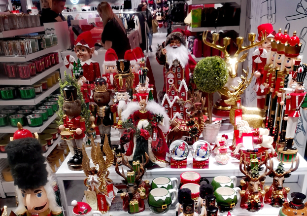 Ladeninneneinrichtung mit Weihnachtsdekorationen, darunter Spielzeuge, Tassen und andere Gegenst├Ąnde auf Tischen und Regalen, mit Menschen, Lichtern und einer Wand im Hintergrund.