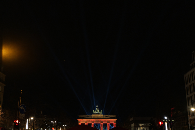 Das Brandenburger Tor in Berlin nachts, beleuchtet von Straßenlaternen, mit Gebäuden, Bäumen, Pfählen, Verkehrszeichen, Schildern und Zelten drumherum gegen einen dunklen Himmel.