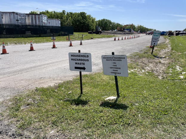 Straßenseite mit "Haushaltsgefährlicher Abfall"-Sammelstelle markiert, mit Fahrzeugen, Gras, Bäumen und einem bewölkten Himmel im Hintergrund.