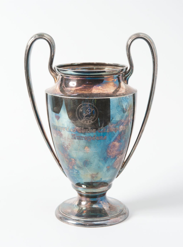 Der UEFA-Champions-League-Pokal, ein stählerner Becher mit zwei Griffen und einem blauen und silbernen Farbschema, der mit "UEFA Champions League" auf einem weißen Hintergrund beschriftet ist.