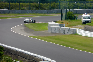 Ein Rennwagen fährt eine Rennstrecke hinunter, im Hintergrund ein Polizeiwagen, umgeben von Gras, Pflanzen, Bäumen und Zaun, mit einem Fahrzeug auf der rechten Seite und Text unten.
