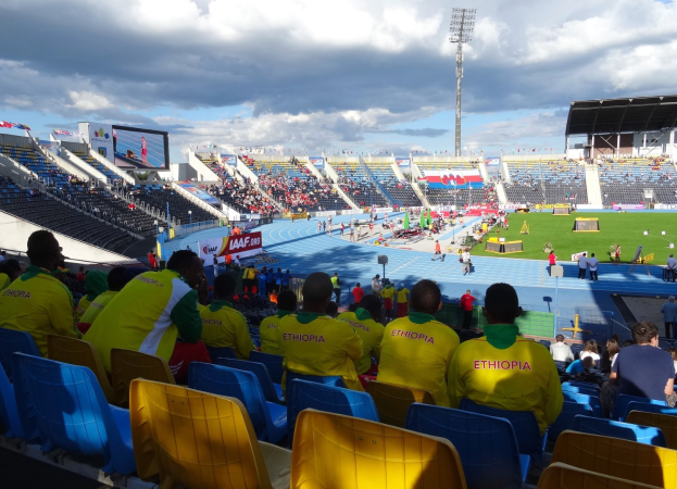 Eine Gruppe von Menschen in einem überfüllten Stadion, das ein Fußballspiel schaut, mit Schildern, einem Anzeigebildschirm und einem bewölkten Himmel.