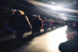 Eine Gruppe von Menschen läuft über einen hell erleuchteten Laufsteg bei einer Modenschau, mit Zuschauern auf beiden Seiten.