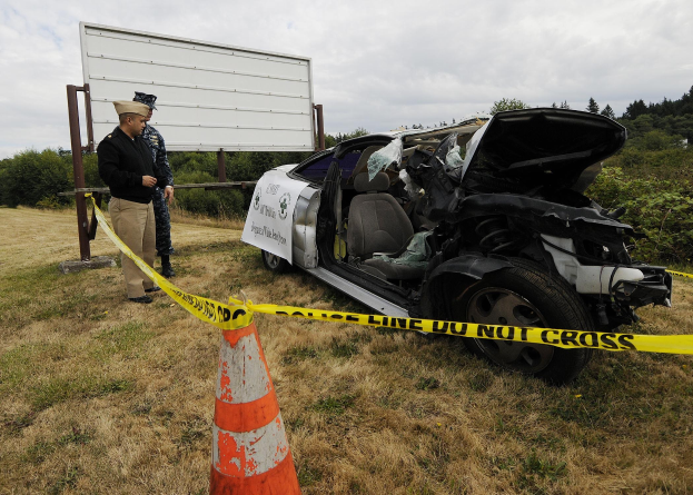 Ein Polizist in Uniform steht neben einem schwerbeschädigten Auto am Straßenrand, mit eingedellter Motorhaube und zersplitterten Fenstern, neben einem Verkehrskegel und einer bandagierten Stange, mit Bäumen und einem bewölkten Himmel im Hintergrund.