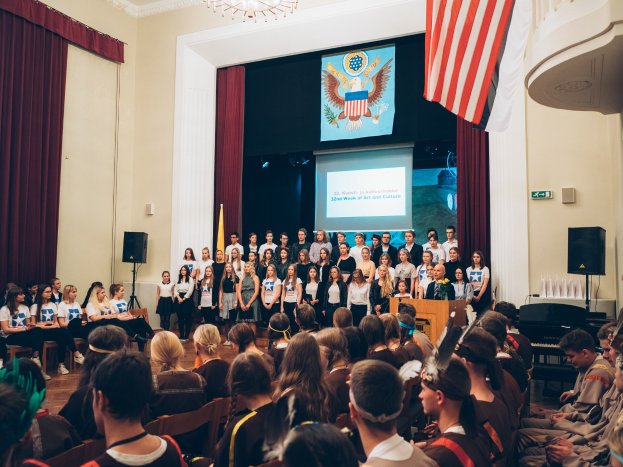 Gruppe von Menschen vor sitzendem Publikum mit Podium, Mikrofon, Flaggen, Lautsprechern, Bildschirm mit Text und Bild, Vorhängen und Kronleuchter während eines Kunst- und Kulturereignisses.