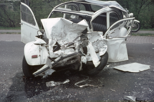 Ein Auto mit schwerer Frontschaden steht am Straßenrand, im Hintergrund sind Bäume, ein Gebäude und ein weiteres Fahrzeug zu sehen.