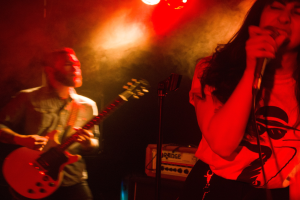 Eine Frau singt in ein Mikrofon auf der Bühne, während ein Mann Gitarre spielt und Deckenlampen das lebendige Ambiente beleuchten.