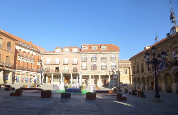 Ein Stadtplatz mit einem zentralen Brunnen, umgeben von Bänken, Topfpflanzen, Straßenlaternen und -masten, mit einem Uhrenturm und Gebäuden im Hintergrund unter einem klaren blauen Himmel.