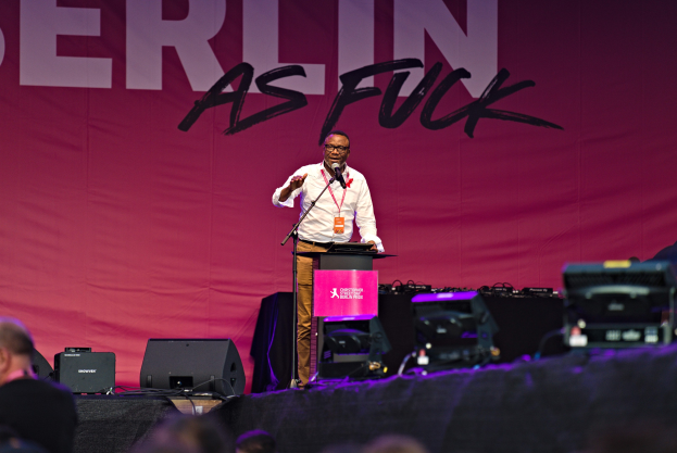 Mann in weißem Hemd und braunen Hosen, der in ein Mikrofon spricht, vor einem pinken "Berlin as Fuck"-Plakat auf der Bühne steht.