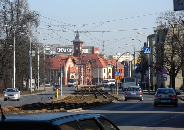 Eine belebte Stadtstraße mit Autos, Bussen, Straßenlaternen, Bahnschienen, Schildern, Laternenmasten, Bäumen, Gebäuden mit Fenstern und einem Uhrenturm unter einem klaren Himmel.
