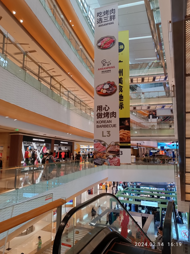 Ein Innenblick in ein Einkaufszentrum mit einer Rolltreppe in der Mitte, Menschen, die herumlaufen, einige mit Handtaschen, und Schilder mit Text, die von der Decke hängen, beleuchtet von Lichtern mit Geschäften im Hintergrund.