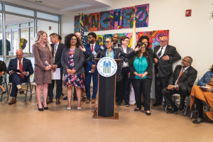 Gruppe von Menschen vor einem Rednerpult mit Mikrofon und einer Tafel mit der Aufschrift "Sancho Stadtrat tritt dem Stadtrat bei", mit sitzenden und stehenden Teilnehmern, Flaggen, Feuerlöscher und Deckenbeleuchtung im Hintergrund.
