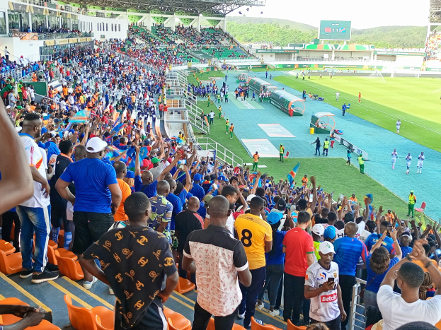 Eine große Menschenmenge versammelt sich vor einem eingezäunten Fußballfeld, einige sitzen und andere stehen, unter einem klaren blauen Himmel mit Hügeln im Hintergrund und warten auf ein Spiel.