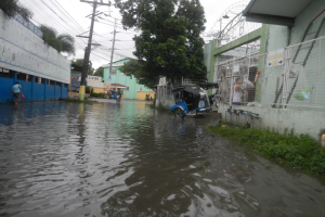 Eine überflutete Straße mit einem geparkten Motorrad in der Mitte, Gebäude, Bäume und Strommasten auf beiden Seiten und einige Menschen und Fahrzeuge im Hintergrund.