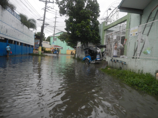 Eine überflutete Straße mit einem geparkten Motorrad in der Mitte, Gebäude, Bäume und Strommasten auf beiden Seiten und einige Menschen und Fahrzeuge im Hintergrund.