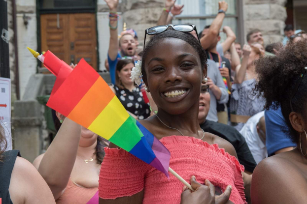 Eine Gruppe von Menschen steht vor einem Gebäude, mit einer Frau in der Mitte, die eine Regenbogenflagge hält, wahrscheinlich bei einem Christopher Street Day; das Gebäude hat Fenster, eine Tür und eine Tafel mit Text.
