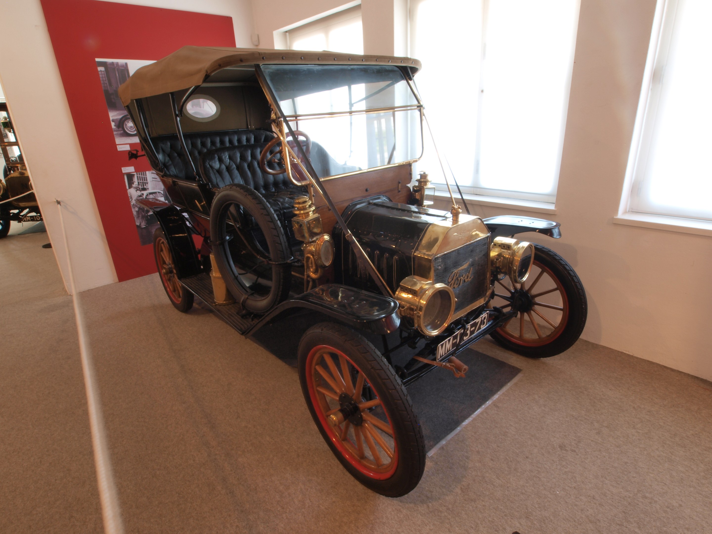 Ein Oldtimer mit glänzendem Lack auf einer Museumsausstellung umgeben von anderen Fahrzeugen, mit einer Informationswand auf der linken Seite und Glasfenstern auf der rechten Seite.