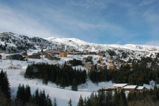 Idyllisches Panorama eines Skigebiets in den französischen Alpen mit schneebedeckten Bergen, Bäumen, Gebäuden und einem klaren blauen Himmel.