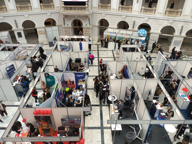 Eine große Gruppe von Menschen versammelt sich in einem Raum voller Stände, Banner, Tische, Stühle und Ballons, mit einem Gebäude im Hintergrund mit Bögen, Säulen, Geländern und einem Bildschirm, was auf eine internationale Fachmesse hinweist.