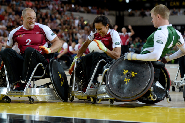 Eine Gruppe von Menschen im Rollstuhl spielt Handball mit einem Ball in der Mitte, mit Zuschauern im Hintergrund, Bild etwas unscharf.
