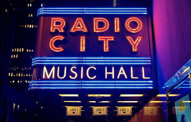 Das erleuchtete Radio City Music Hall in New York City mit hohen Gebäuden im Hintergrund, das eine 'Radio City' Leuchtschrift und zusätzliche Schilder auf der rechten Seite zeigt.