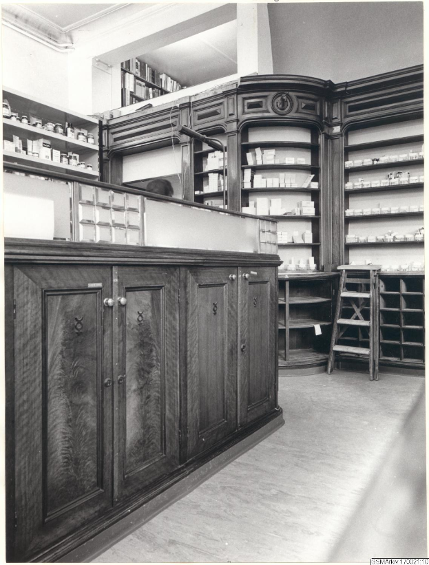 Schwarz-weiß-Foto eines vintage Apothekeninneren mit einer Theke, Schränken, einer Leiter und Regalen, die mit verschiedenen Gegenständen gefüllt sind, mit Text oben.