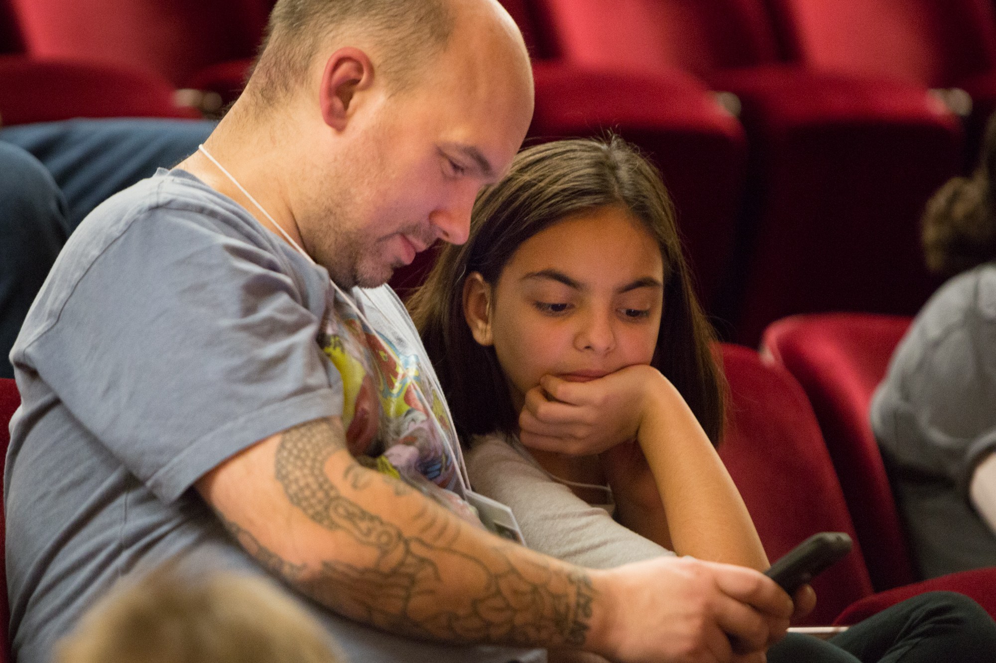 Ein Mann und ein junges Mädchen sitzen in einem Kino und schauen beide auf ein Handy, wobei der Mann es hält und das Mädchen es aufmerksam betrachtet.