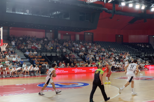 Eine Gruppe von Menschen, die Basketball in einer Turnhalle spielen, mit einem Basketballkorb auf der linken Seite und Zuschauern auf Stühlen im Hintergrund, unter hellen Stadionlichtern.