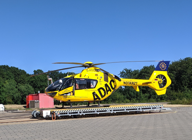 Gelber Hubschrauber auf einem Flughafenvorfeld mit umliegenden Bäumen und klarem blauen Himmel, wahrscheinlich für Luftrettungsdienste in Deutschland verwendet.